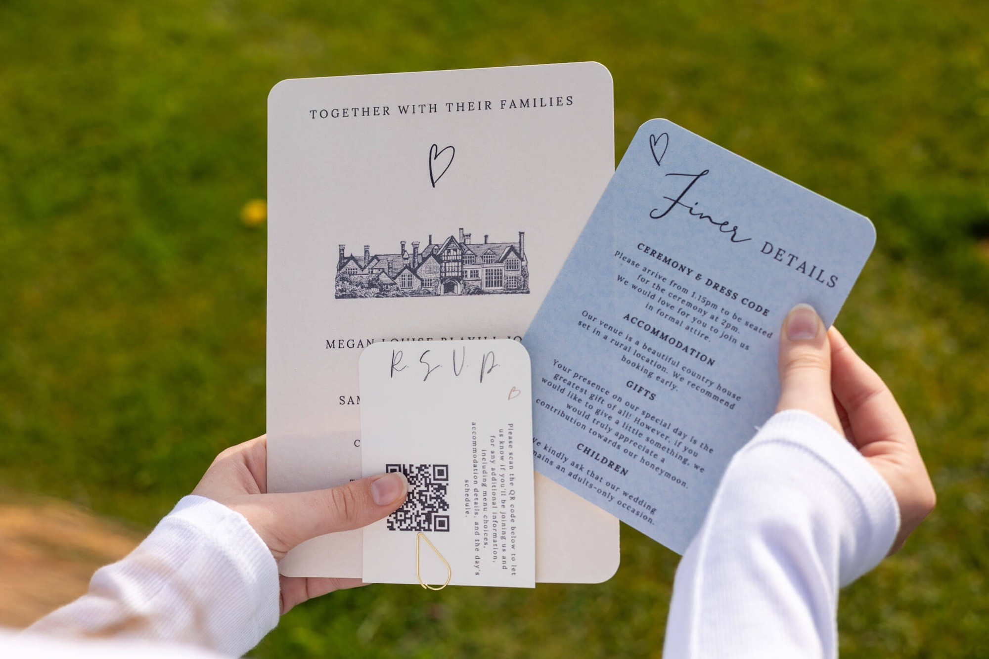 A person holds three wedding invitation cards, including the main invite, an RSVP card with a QR code, and a blue details card, all against a grassy outdoor background.