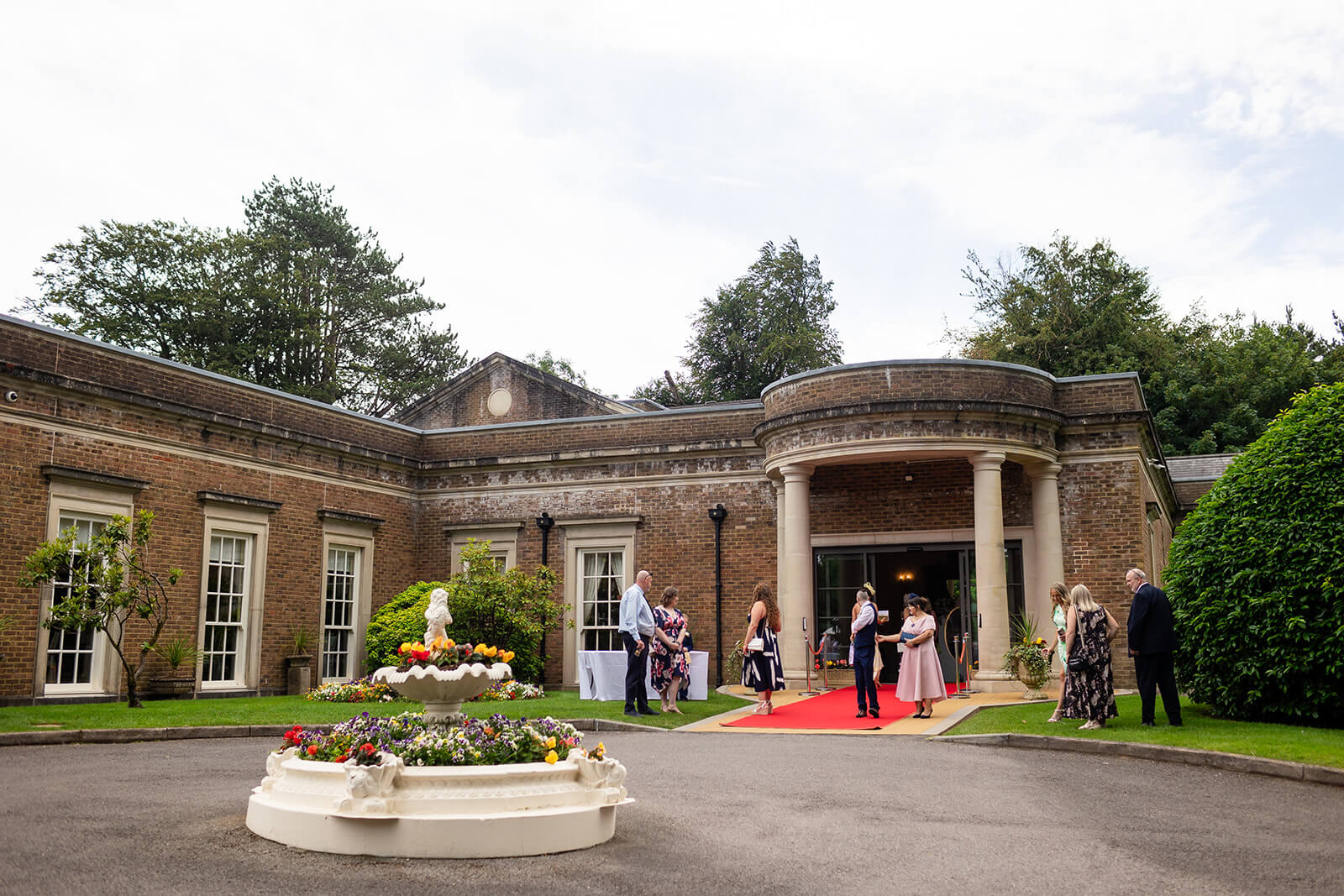 People gather outside a brick building with columns and a red carpet, surrounded by greenery and a round flower fountain in front.